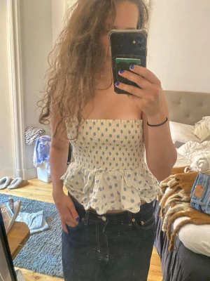 Vit prickig bandeau topp med volang - Supersöt bandeau topp i vitt med ljusblå prickar och volangkant nedtill. Smockad överdel som sitter tight och ger en feminin siluett. Perfekt för varma dagar och enkel att styla med jeans eller kjol.