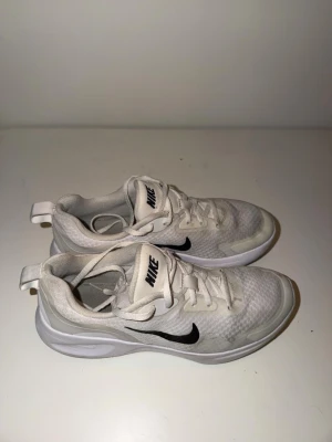 Vita Nike sneakers med svart swoosh - Fräscha vita sneakers från Nike med svart swoosh på sidan. Skorna har en sportig design med mesh-ovandel och vita snören. Perfekta för dig som gillar en clean och stilren look. Lätta och bekväma med låg profil.
