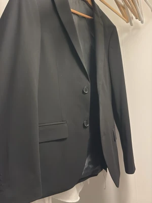 Svart klassisk kavaj med två knappar - Stilren svart kavaj med två knappar framtill och klassiska slag. Kavajen har raka fickor med lock och är helfodrad. Perfekt för en dressad look och passar till många olika outfits. Kom med förslag på pris, kostar runt 2,500 ny, och den är ny.