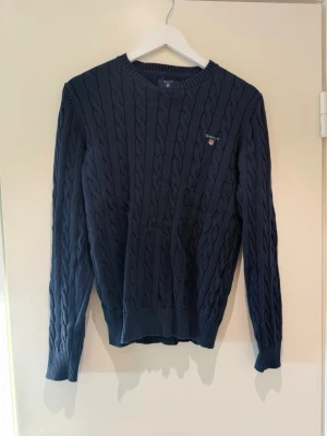 Mörkblå kabelstickad tröja Gant - Mörkblå kabelstickad tröja från Gant i premium bomull. Tröjan har rund halsringning, ribbade muddar och Gant-logga broderad på bröstet. Klassisk och stilren design som passar perfekt till jeans eller chinos.