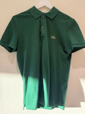 Grön pikétröja från Lacoste - Klassisk grön pikétröja från Lacoste med korta ärmar och krage. Tröjan har två knappar framtill och den ikoniska krokodilloggan broderad på bröstet. Slim fit-modell i bomull som passar perfekt till jeans eller shorts.