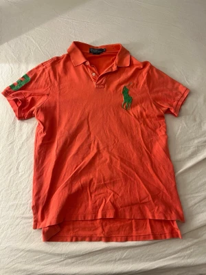 Orange pikétröja från Ralph Lauren - Snygg orange pikétröja från Ralph Lauren med klassisk krage och knappar. Stor grön logga på bröstet och en grön siffra 3 på ärmen. Perfekt för en sportig och avslappnad stil.