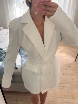 Vit oversized kavaj med fickor och figursydd - Stilren vit kavaj med oversized passform och markerade axlar. Kavajen har breda slag, två stora fickor framtill och knäppning med tre knappar. Materialet ser ut att vara ett slätt tyg, troligen polyesterblandning. Perfekt för en trendig och modern look.