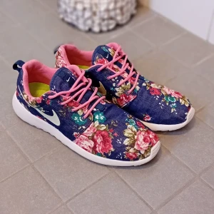 Blommiga Nike sneakers med rosa detaljer - Unika sneakers från Nike med mörkblå bas och färgglatt blommönster. Skorna har rosa snören, rosa innerfoder och vit sula. Ikonisk vit Nike-logga på sidan. Perfekta för dig som vill sticka ut med en färgglad och trendig look.