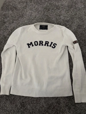 Vit stickad tröja från Morris - Vit stickad tröja från Morris med svart textlogga på bröstet och Union Jack-detalj på ärmen. Tröjan har rund halsringning, långa ärmar och ribbade muddar vid ärmslut och nederkant. Perfekt för en stilren och avslappnad look.