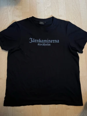 Svart t-shirt Järnkaminerna  - Säljer en svart t-shirt från South West med trycket 'Järnkaminerna Stockholm' i vitt på bröstet. Tillverkad i ekologisk bomull och har klassisk passform med korta ärmar och rund hals. Perfekt för dig som vill visa ditt stöd för Järnkaminerna.