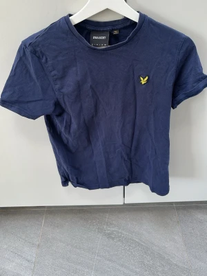 Marinblå t-shirt från Lyle & Scott - Snygg marinblå t-shirt från Lyle & Scott med klassisk gul logga på bröstet. T-shirten har rund halsringning och korta ärmar, tillverkad i mjuk bomull för skön känsla. Perfekt till jeans eller shorts för en clean och enkel stil.