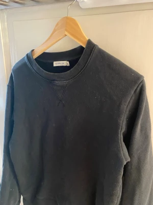 Svart sweatshirt från tiger of Sweden  - En klassisk svart sweatshirt från tiger of Sweden med rund halsringning och ribbade muddar. Tröjan har en liten gul detalj nertill och är tillverkad i mjukt material, perfekt för en avslappnad stil.