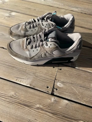Nike Air Max 90 grå sneakers - Säljer ett par grå Nike Air Max 90 sneakers med klassisk siluett. Skorna har mesh och mockadetaljer, samt vit och svart sula med synlig Air-enhet. Swoosh-logga på sidan och Air Max-märke i turkos på hälen. Perfekta för dig som gillar streetwear.