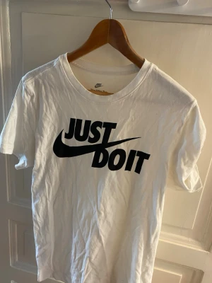 Vit Nike t-shirt med Just Do It-tryck - Snygg vit t-shirt från Nike med det klassiska svarta 'Just Do It'-trycket på bröstet. T-shirten har rund hals och korta ärmar, perfekt för en sportig och avslappnad stil. Tillverkad i mjuk bomull för skön komfort.