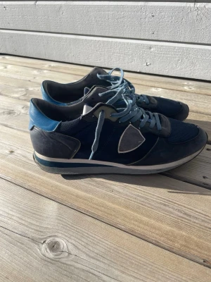 Mörkblå sneakers från Philippe Model - Snygga mörkblå sneakers från Philippe Model med ljusblå detaljer och vita inslag. Skorna har mesh och läderpaneler, samt en ikonisk sköldlogga på sidan. Ljusblå snörning och bekväm sula med kontrastfärg. Perfekta för en trendig streetstil.