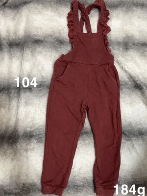 Vinröd byxdress med volangband - Byxdress med vinröd färg, volangprydda axelband och ribbade benslut. Två fickor framtill och relaxed passform ger en chill vibe. Perfekt för dig som gillar bekväma och snygga plagg.🌸 Fint använt skick. Storlek 104