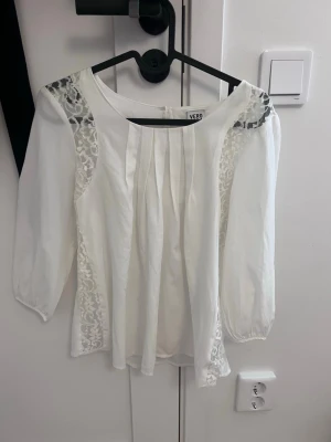 Vit blus med spets från Vero Moda - Snygg vit blus från Vero Moda med trekvartsärm och fina spetsdetaljer på axlar och sidor. Blusen har veck framtill och en lös, luftig passform. Perfekt att styla med jeans eller kjol för en trendig look.