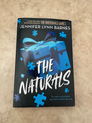 The Naturals av Jennifer Lynn Barnes - En spännande ungdomsroman på engelska med svart och blått omslag, dekorerat med pusselbitar och ett paket med blå rosett. Perfekt för dig som gillar mysterier och thrillers.