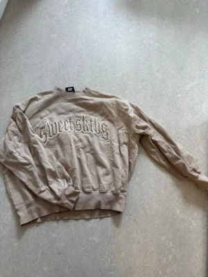 Beige sweatshirt från Sweet SKTBS - Säljer en beige sweatshirt från Sweet SKTBS med tryckt logga på bröstet. Tröjan har rund hals, ribbade muddar och avslappnad passform. Perfekt för en streetwear-look och enkel att matcha med jeans eller cargo.