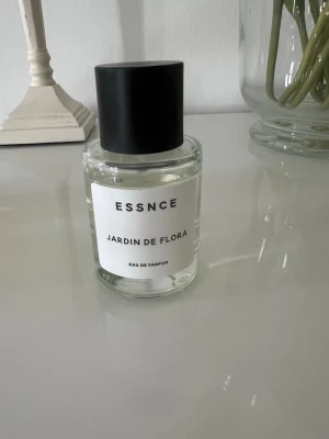 ESSNCE Jardin de Flora parfym - Fräsch parfym från ESSNCE med namnet Jardin de Flora. Flaskan är genomskinlig med en svart, rund kork och vit etikett. Eau de Parfum med stilren och modern design, perfekt för dig som gillar blommiga dofter.