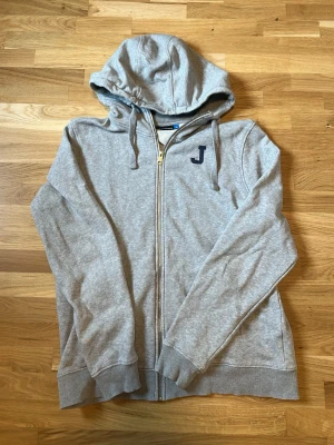 Grå hoodie J.lindeberg - Grå hoodie med dragkedja från J.Lindeberg. Mjuk bomullsblandning, huva med dragsko och ribbade muddar. Svart J-logga på bröstet ger en clean look. Perfekt till jeans eller joggers.