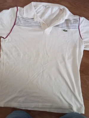 Vit pikétröja från Lacoste - Vit pikétröja från Lacoste med klassisk krage, knäppning framtill och tunna marinblå ränder över bröstet. Tröjan har röda detaljer längs axlarna och det ikoniska gröna krokodilmärket på vänster sida. Perfekt för en sportig och stilren look. Jag har storlek s också och M
