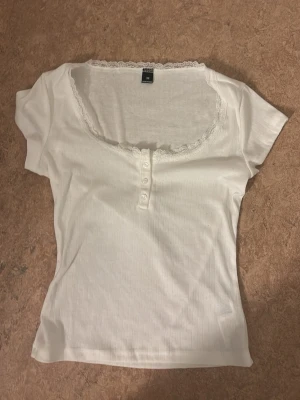 Vit topp med spets och knappar - Söt vit topp med korta ärmar, spetskant vid halsringningen och dekorativa knappar framtill. Tunn och mjuk i materialet, perfekt för en enkel och stilren look. Figurnära passform och lätt ribbad struktur.