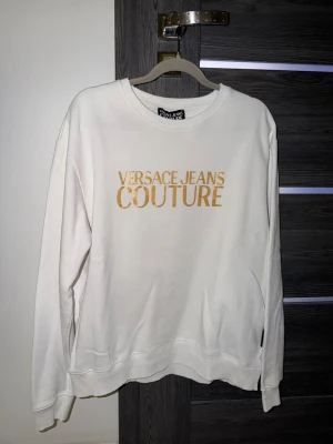 Vit sweatshirt Versace Jeans Couture - Vit sweatshirt med guldig logotyp från Versace Jeans Couture. Tröjan har rund halsringning, långa ärmar och ribbade muddar vid ärmslut och nederkant. Perfekt för en trendig och avslappnad stil.