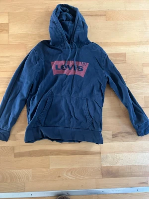 Blå hoodie från Levi's med logga - Mörkblå hoodie från Levi's med stor röd logga på bröstet. Klassisk känguruficka framtill och justerbar huva med dragsko. Tillverkad i mjukt bomullsmaterial, perfekt för en avslappnad och cool stil.