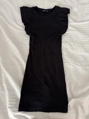 Svart kort klänning med volangärm - En svart, ribbad kort klänning från Zara med volangdetaljer på ärmarna. Klänningen har en rund halsringning och är figurnära i modellen. Perfekt för dig som gillar en enkel men ändå trendig look.