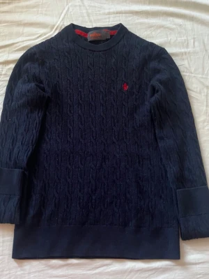 Mörkblå kabelstickad tröja Morris barnstorlek 150 - Kabelstickad mörkblå tröja från Morris med ett rött broderat emblem på bröstet. Perfekt för en stilren och klassisk look. Krymt i tvätten från S till barnstorlek 150