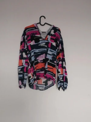 Färgglad mönstrad blus med V-ringning - Blus med abstrakt mönster i rosa, orange, svart, blått och vitt. V-ringad framtill och rundad nederkant. Långa ärmar med resår vid handlederna. Perfekt för dig som gillar färg och unika plagg. Materialet känns lätt och luftigt.