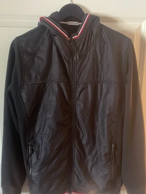 Svart Moncler vindjacka med huva - Svart vindjacka/cardigan från Moncler med huva och dragkedja framtill. Jackan har röda och vita detaljer vid krage och nederkant samt två fickor med dragkedja. Moncler-logga på ärmen. Perfekt för dig som vill ha en stilren och sportig look.