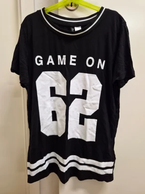 Svart t-shirt med GAME ON-tryck XS H&M  - Cool svart t-shirt med vit text 'GAME ON' och siffran 62 framtill. T-shirten har sportig känsla med vita ränder vid nederkant och halsringning. Skön passform och tillverkad i mjuk bomull, perfekt för en avslappnad streetstyle.