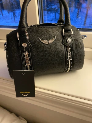 Svart handväska Zadig & Voltaire - Cool svart handväska i strukturerat läder från Zadig & Voltaire. Väskan har silverfärgade detaljer, dubbla handtag, dekorativa dragkedjor och nitar samt en ikonisk vinge framtill. Perfekt för dig som gillar edgy och trendig stil. 21 cm lång 