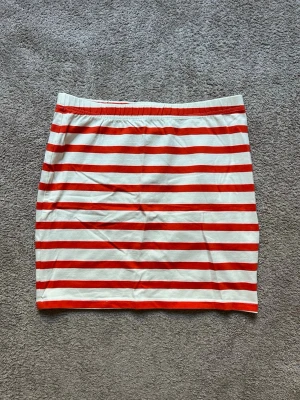 Randig kortkjol från Holly & Whyte - Kort kjol från Holly & Whyte by Lindex. Kjolen har resår i midjan och är i 95% bomull och 5% elestan. Så den har en viss stretch i sig. Ränderna är vita och röda (lite åt det orangea hållet) . Kjolen är ny och oanvänd, men utan prislapp kvar. 