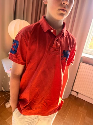 Röd pikétröja från Polo Ralph Lauren - Snygg röd pikétröja från Polo Ralph Lauren med klassisk krage och knappar framtill. Tröjan har en stor blå logga med ryttare på bröstet och siffran 3 i blått på ena ärmen. Tillverkad i bomull och har korta ärmar.