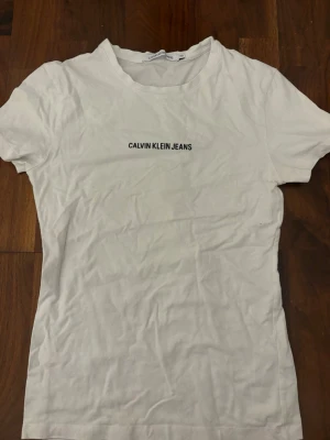 Vit t-shirt från Calvin Klein Jeans - Vit t-shirt från Calvin Klein Jeans med svart logotyptryck på bröstet. Klassisk rund halsringning och korta ärmar. Gjord i mjuk bomull för en bekväm känsla.