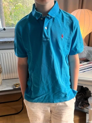 Blå pikétröja från Ralph Lauren - Snygg blå pikétröja från Ralph Lauren med klassisk krage och korta ärmar. Tröjan har två knappar vid halsen och den ikoniska röda loggan broderad på bröstet. Perfekt till jeans eller shorts för en avslappnad stil.