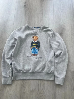Grå sweatshirt Polo Bear Ralph Lauren - Grå sweatshirt från Ralph Lauren med ikoniskt Polo Bear-tryck på bröstet. Tröjan har rund halsringning och ribbade muddar vid ärmslut och nederkant. Perfekt för en avslappnad och trendig stil.