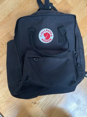 Svart Fjällräven Kånken ryggsäck - Klassisk svart Fjällräven Kånken ryggsäck i slitstarkt material med det ikoniska runda röda och vita märket framtill. Ryggsäcken har ett stort huvudfack, en framficka med dragkedja och två sidofickor. Justerbara axelband och bärhandtag upptill.