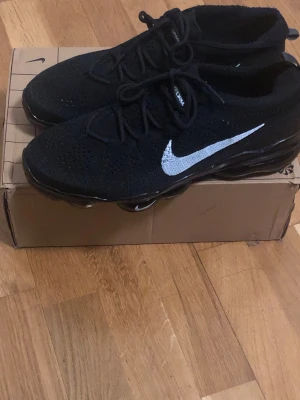 Nike vapormax  - Snygga svarta Nike Flyknit sneakers med vit swoosh på sidan. Skorna har en stickad ovandel som ger en lätt och luftig känsla, samt klassisk snörning. Perfekta för dig som gillar sportig och stilren look.