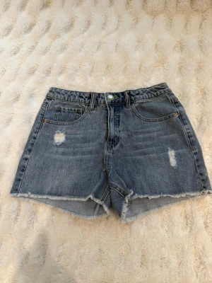 Blå denimshorts med fransig kant - Säljer ett par blå denimshorts med fransig rå kant och slitna detaljer framtill. Klassisk femficksmodell med knapp och dragkedja. Perfekta för sommaren och ger en avslappnad look.