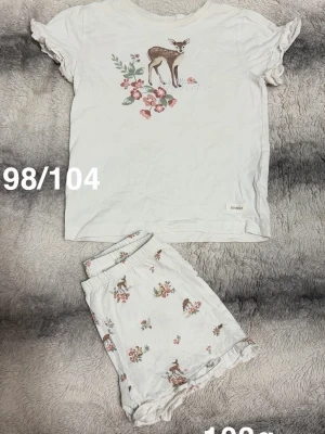 Vit kortärmad pyjamas - Söt vit pyjamas från Newbie med kortärmad topp och shorts. Båda delarna har gulligt djurtryck och blommönster. Toppens ärmar och shortsen har volangdetaljer. Perfekt för mysiga kvällar och morgnar.🌸 Fint använt skick. Storlek 98/104