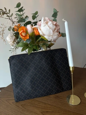 Brun datorväska från By Malene Birger - Snygg datorväska i brunt med By Malene Birgers klassiska mönster. Väskan har guldfärgad dragkedja och är tillverkad i tåligt material med beige insida. Perfekt för att bära din laptop med stil. Se mått på bild 