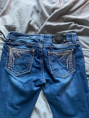 Blå Miss Me jeans med strassdetaljer - Snygga lågmidjade  blå jeans från Miss Me med vita kontrastsömmar och blingade bakfickor med strass och broderier. Klassisk femficksmodell och låg midja. Perfekta för dig som vill sticka ut med detaljer och cool stil.