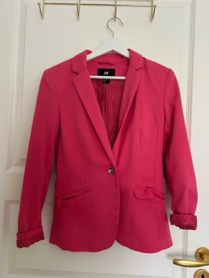 Rosa kavaj från H&M med knapp - Snygg rosa kavaj från H&M med klassisk krage, en knapp framtill och två fickor med lock. Kavajen har ett slätt tyg och fodrade ärmar med diskret randigt foder som syns när du viker upp ärmsluten. Perfekt för att addera färg till din outfit.