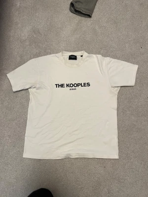 Vit t-shirt från The Kooples - Vit t-shirt från The Kooples med svart tryck på bröstet där det står 'THE KOOPLES PARIS'. Klassisk rund halsringning och korta ärmar. Tillverkad i mjuk bomull för en skön känsla och enkel stil.
