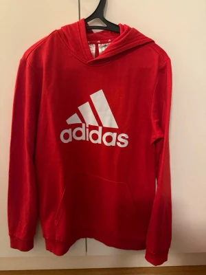 Röd Adidas hoodie med logga - Riktigt snygg röd hoodie från Adidas med stor vit logga på bröstet. Klassisk modell med huva och känguruficka framtill. Perfekt för dig som gillar sportig och avslappnad stil. Mjuk och skön bomullsblandning som passar året runt.