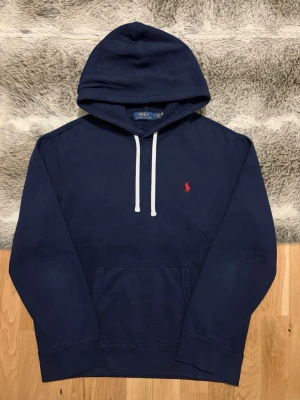 Marinblå Polo Ralph Lauren Fleece Hoodie - Storlek L - Hej! 👋 Säljer en riktigt snygg marinblå Polo Ralph Lauren hoodie i storlek L med röd pony-logga. 🐎 Tröjan är i mycket gott skick, välvårdad och utan defekter. Garanterat äkta med alla originaltvättlappar kvar. QR-kod finns på lappen men visar för tillfället ”authentication check” på grund av Ralph Laurens säkerhetsspärr (antagligen skannad för mycket tidigare). Ett klockrent premium-basplagg till garderoben! Fraktar snabbt! 💬 100% 🚛📦 