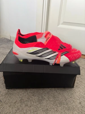 Adidas Predator fotbollsskor röd/vit - Snygga Adidas Predator fotbollsskor i rött, vitt och svart med gula detaljer. Skorna har låg profil, klassisk tre-randig design och textilöverdel med gummidetaljer för extra grepp. Yttersulan är gjord för fasta underlag och har mönstrade dobbar för bra fäste. Helt nya storlek 43 1/3