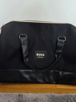 Svart weekendbag från BOSS Parfums - Snygg svart weekendbag från BOSS Parfums med stilren design. Väskan har dubbla handtag i konstläder, detaljer i silver och en praktisk dragkedja upptill och sidofack för extra förvaring. Köpt från Boozt.com och aldrig använd.