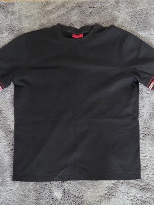 Hugo T-Shirt - Svart t-shirt från Hugo med korta ärmar och snygga röda och vita logodetaljer på ärmsluten. T-shirten har en klassisk passform och är gjord i mjuk bomull, perfekt för en stilren och modern look. 