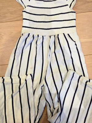 Randig jumpsuit från Mywear - Säljer en vit jumpsuit från Mywear med svarta horisontella och vertikala ränder. Jumpsuiten har korta ärmar, smockad överdel och vida ben. Perfekt för en avslappnad och trendig look i sommar.